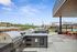 Modern rooftop space with built-in stainless steel grills, dining tables, red chairs, and skyline views in the background.