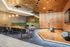 Modern lounge area with green velvet banquette seating, mixed tables and chairs, a wooden stage with microphones, and a metallic gold ceiling.