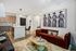 Modern apartment living room with a velvet red sofa, patterned rug, coffee table, kitchen in background, and black-and-white wall art of four women.