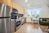 Modern kitchen with stainless steel appliances, light wood cabinetry, a small white desk under a large window, green sofa, and hardwood floors.
