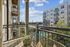Apartment balcony with modern railing and a lounge chair, offering views of a pool, courtyard, and neighboring buildings under a bright blue sky.