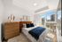 Bright and modern bedroom with chic decor, featuring a plush bed, wooden dresser, and large windows with a stunning urban view.