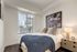 Elegant bedroom with a cozy bed, large window showcasing city views, stylish artwork, and modern bedside table with a chic lamp.
