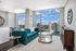 Bright and stylish living room featuring a teal velvet sofa, elegant glass coffee table, and expansive city views through floor-to-ceiling windows.