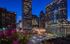 Vibrant cityscape view from an apartment balcony featuring blooming flowers, illuminated skyscrapers, and a bustling downtown ambiance at dusk.