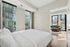 Bright, modern apartment bedroom with luxurious bedding and floor-to-ceiling windows showcasing stunning city views, leading to a cozy sitting area.