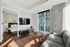 Modern apartment living room with a sleek grey sofa, wall-mounted TV, and large windows offering city views, adjacent to a cozy bedroom.