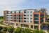 Modern multi-story apartment building with red brick facade, large windows, and inviting balconies set amidst lush greenery.