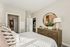 Bright and inviting bedroom with stylish decor, featuring a large mirror above a wooden dresser, soft bedding, and a glimpse into an adjoining room.