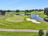 Scenic golf course view featuring lush greens, a tranquil pond, and a modern clubhouse under a clear blue sky.
