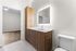Bright and modern bathroom featuring a sleek wooden vanity with ample storage, an illuminated mirror, and contemporary fixtures, leading to an adjacent room with elegant wooden flooring.