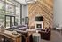 Luxurious living room with high ceilings, elegant chandelier, and large windows overlooking a verdant garden. Features cozy leather seating, rustic wood accent wall, and stylish fireplace beneath a modern TV.