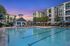 Luxurious apartment complex with a large pool, surrounded by palm trees and stylish lounge chairs, under a vibrant sunset sky.