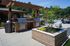 Outdoor luxury grilling area with modern stainless steel barbecues, surrounded by lush greenery and a resort-style swimming pool.