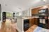 Modern open-concept kitchen and living area with sleek wooden cabinetry, granite countertops, and vibrant decor, leading to a bright seating area with large windows overlooking greenery.