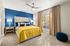 Bright, elegant bedroom featuring a plush bed with vibrant yellow bedding, blue accent wall, and large sunlit window, perfect for a cozy and inviting atmosphere.