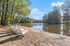 Tranquil lakeside scene featuring white Adirondack chairs under tall trees, perfect for relaxing and enjoying nature's beauty.