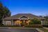 Charming residential building with elegant exterior lighting, surrounded by lush greenery and vibrant flowers at dusk.