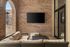 Chic covered patio featuring a cozy wicker sofa set, mounted flat-screen TV on rustic brick wall, and elegant glass doors.