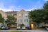 Charming three-story apartment building with elegant balconies, surrounded by lush trees and a serene parking area underneath a bright blue sky.