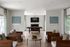 Elegant living room with contemporary wooden furniture, stylish stone fireplace, and artistic decor on white-paneled walls. Large windows offer abundant natural light complemented by soft green curtains.