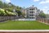 Charming apartment complex with a vibrant green lawn, adjacent to a sleek blue tennis court, surrounded by lush greenery and modern architecture.