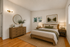 Elegant bedroom with natural wooden furniture, featuring a cozy bed, stylish dresser, and serene artwork. Soft lighting and a large window create a warm, inviting ambiance.