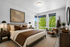 Elegant bedroom with plush bedding, warm leather accents, and large windows offering serene views of lush greenery.