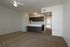 A spacious living room with tan carpeted floor. The kitchen is located at the back of the room.
