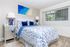 A white bedsheet with blue accents is spread across a bed, accompanied by a blue lamp shade. In the background, a window and ocean-themed artwork add a coastal touch