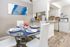 A dining room table set with blue placemats and white plates featuring blue accents, positioned against wall art. The scene includes a view into a white kitchen with a microwave nook