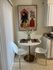 A small dining area features a round table with two white chairs. A vase with flowers sits on the table. A framed abstract artwork hangs on the wall behind. Vertical blinds are partially open.