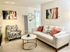 A cozy living room with a patterned armchair, a white sofa with colorful pillows, a round coffee table, and abstract wall art. A floor lamp and a hexagonal shelf adorn the space.
