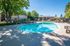 Outdoor pool area with lounge chairs and umbrellas surrounded by a fence and trees, set in a residential community under a clear blue sky.