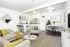 A modern open-plan living space with a beige sofa, round coffee table, dining area, and kitchen. Decor includes yellow accents, a wall mirror, and a ceiling fan. Black bar stools at the counter.
