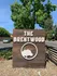 A wooden sign reading "The Brentwood" with a tree emblem below. Surrounded by greenery and stones, under a clear blue sky. Calm, welcoming setting.