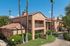 Apartment complex with beige stucco walls and terracotta roof, surrounded by tall palm trees and greenery. Bright, clear blue sky overhead.