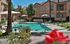 A sunlit outdoor pool area with lounge chairs and umbrellas; surrounded by a peach-colored building and palm trees, conveying a relaxed, vacation vibe.