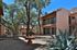 Sunny courtyard of a Mediterranean-style apartment complex with two-story buildings, stairs, trees, and a central terracotta birdbath, flanked by succulents.