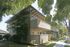 Two-story apartment complex with wooden balconies, surrounded by lush green trees and manicured lawn. Sunlight filters through leaves, creating a serene atmosphere.