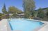 Sunny outdoor scene of a swimming pool surrounded by trees and lounge chairs. Colorful beach balls float on the water, creating a relaxing atmosphere.