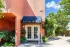 Entrance to a rental office with a blue awning and glass doors, surrounded by lush greenery. The building is peach-colored under a clear blue sky.