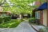 A tranquil courtyard with shade from lush green trees, surrounded by buildings with beige doors and blue awnings. A curved path and manicured lawn create a peaceful atmosphere.
