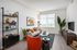 A modern, cozy living room with a white sofa, orange chair, and plants. There's a window with blinds, art on the wall, and a TV on the right.