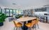 Modern lounge space with light wood floors, featuring a wooden table surrounded by orange chairs. Green sofa and plants add a cozy vibe.