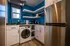 Compact modern kitchen with white cabinets, blue walls, and wood flooring. Features a washing machine, stainless steel fridge, and open shelving with appliances.