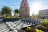 Rooftop terrace with giant chessboard surrounded by planters and trees, overlooking a tall building. Sunlight creates a bright, serene ambiance.