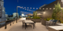 Rooftop at dusk with string lights over a long table and chairs. Grill and greenery on the right, city skyline visible in the background, serene atmosphere.
