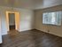 Empty room with light brown hardwood floor and cream walls. A large window with blinds lets in sunlight. Open doorway to another room. Calm ambiance.