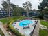 A landscaped apartment complex courtyard with a bright blue swimming pool and hot tub surrounded by lounge chairs, grassy areas, mature trees, and dark blue and gray multi-story buildings.